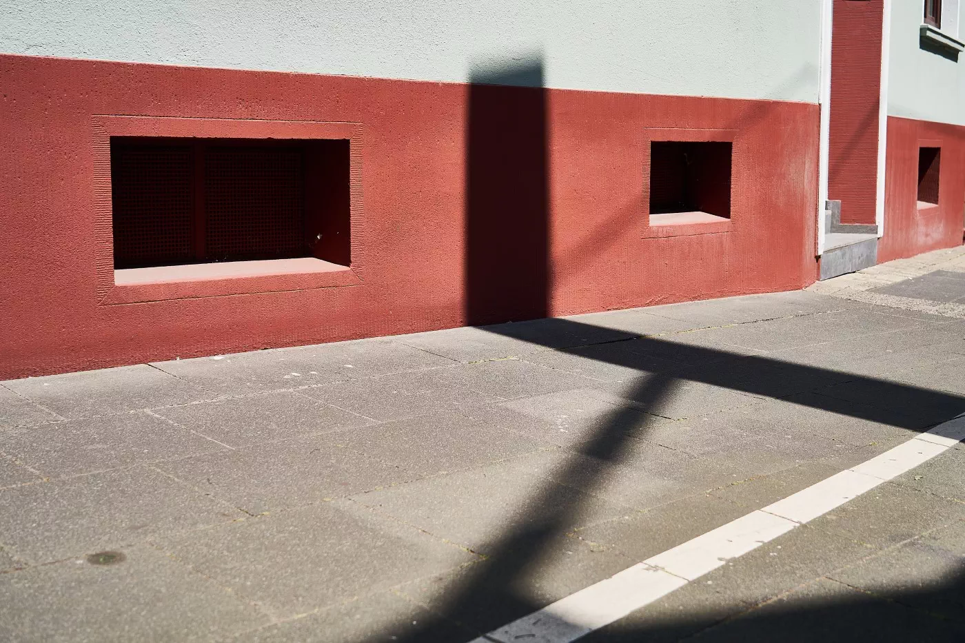 Auf einem von der hellen Sonne beschienenen und mit Betonplatten gepflasterten Bürgersteig sind vor einer rot gestrichenen Hauswand schwarze Schattenlinien zu sehen, die sich auf der Hauswand fortsetzen.