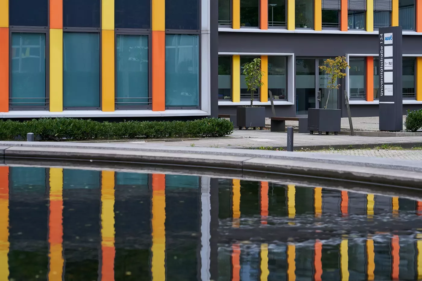 An office building with vertical color stripes in yellow, orange and red hues that is reflected in a pond in front of it.
